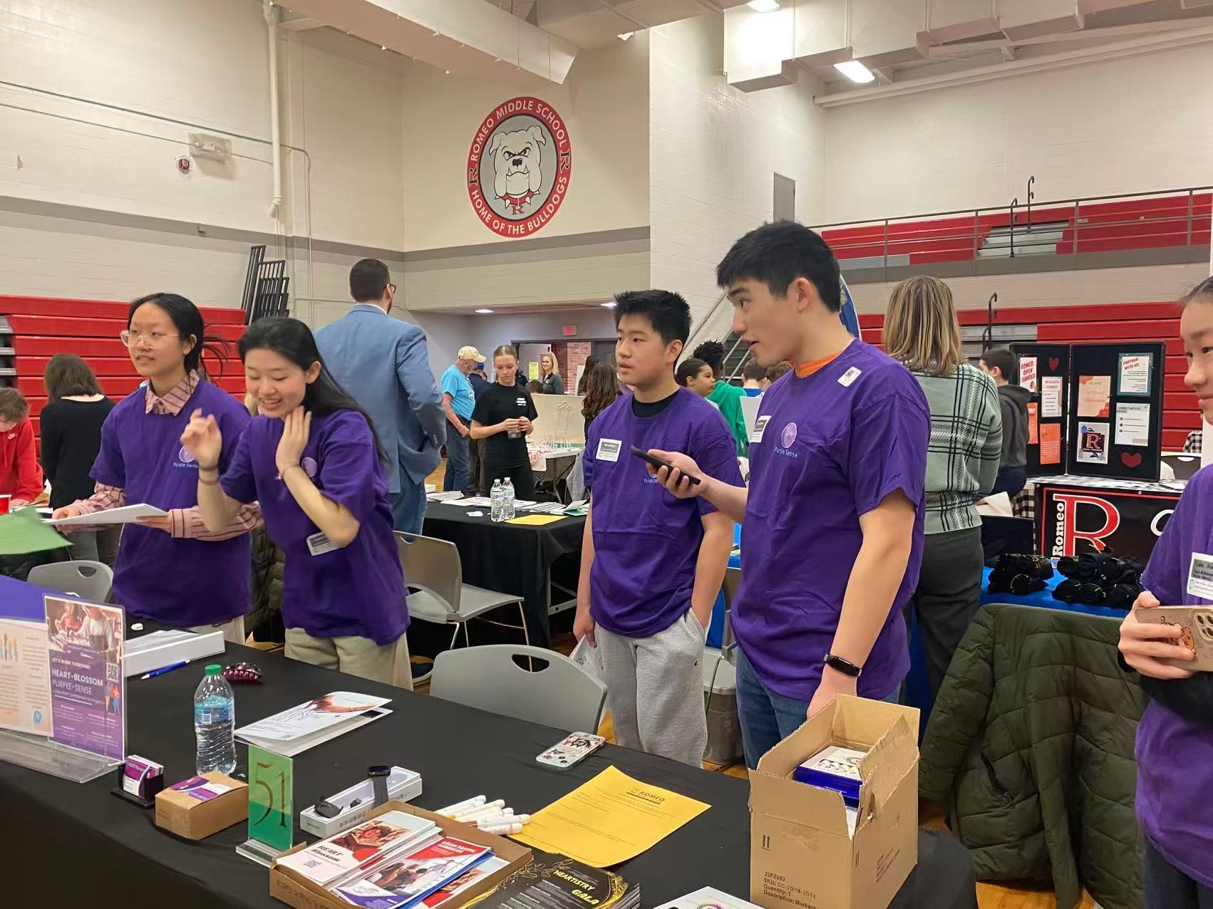 Students in purple shirts at a booth, possibly a science fair. Red and white gym setting.