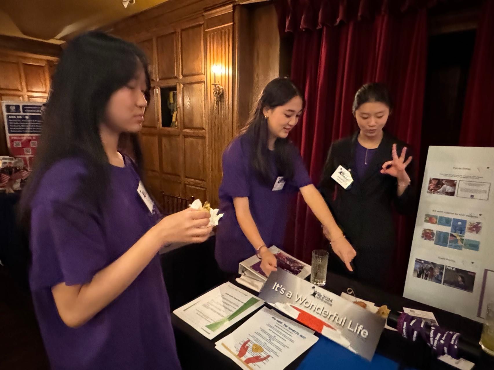 Three Asian women at a table with a display. They wear purple shirts or a suit, gesturing and pointing.