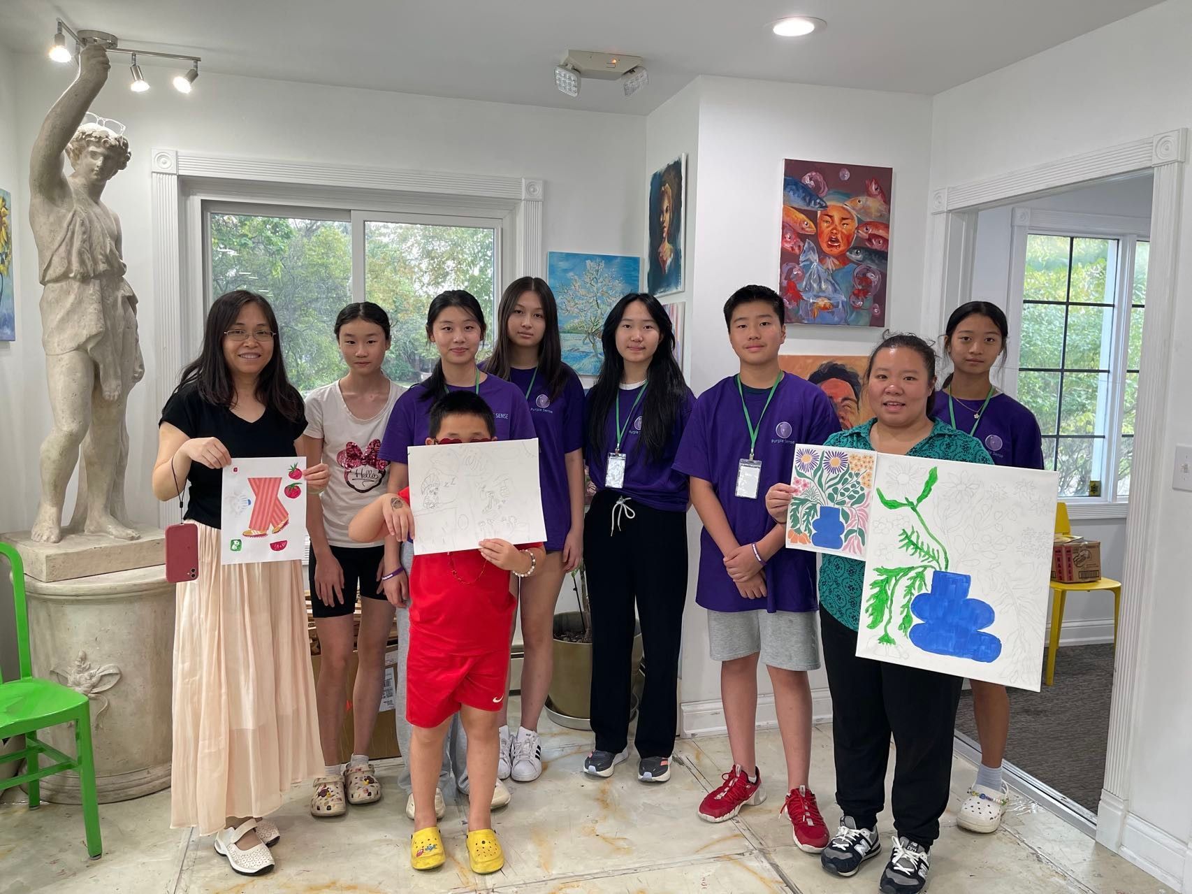 Group of students in art studio holding up paintings. Includes a statue and artwork on the wall.