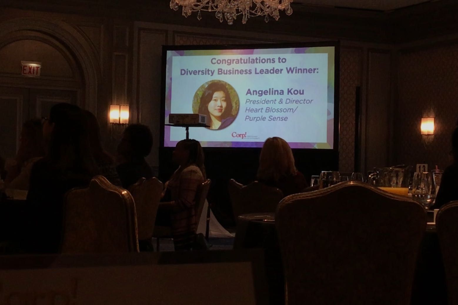 Angelina Ko, Diversity Business Leader winner, on a screen at an event. People seated at tables, dimly lit room.