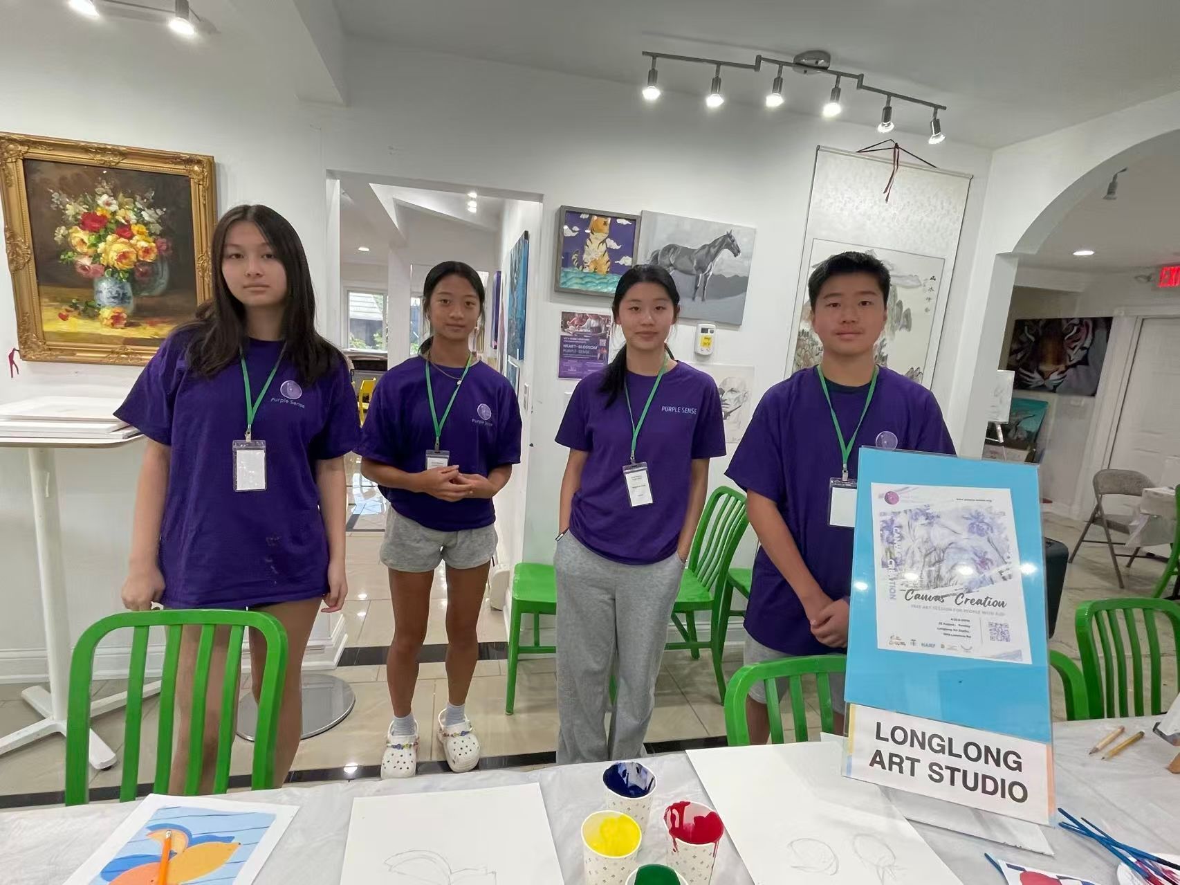 Four young people in purple shirts at an art studio; they stand behind a table with art supplies and paintings.