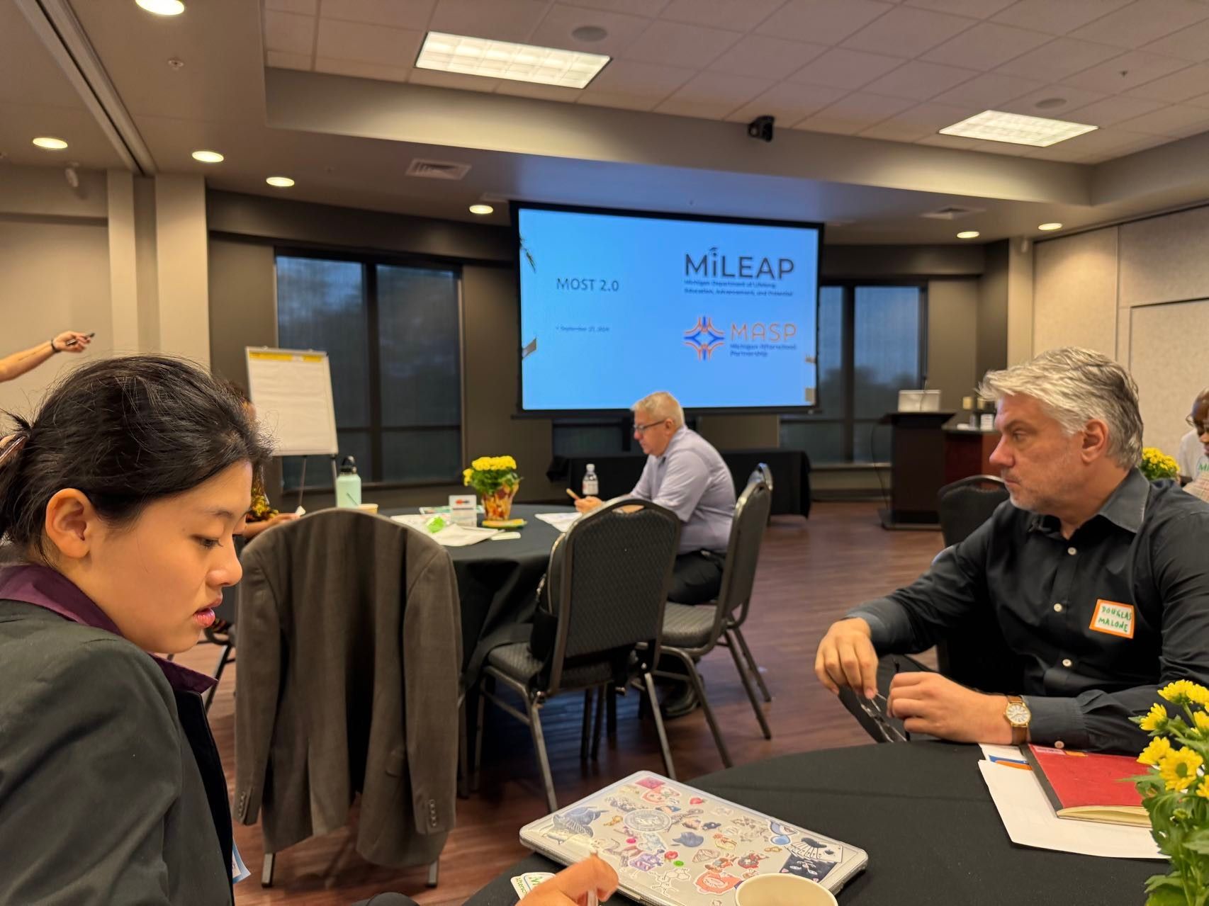 People at round tables in a meeting room, looking at papers and a laptop. A presentation screen is visible.