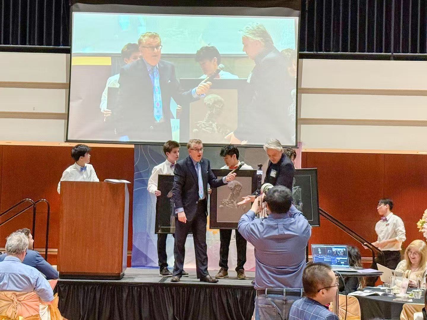 People on a stage at an event; men in suits are handing a framed award to younger people.