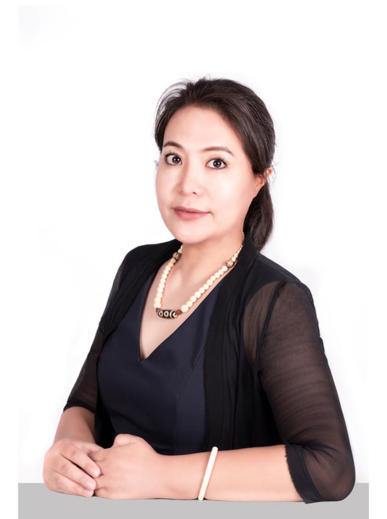 Woman with dark hair, wearing a necklace and black jacket, seated with hands clasped, smiling.