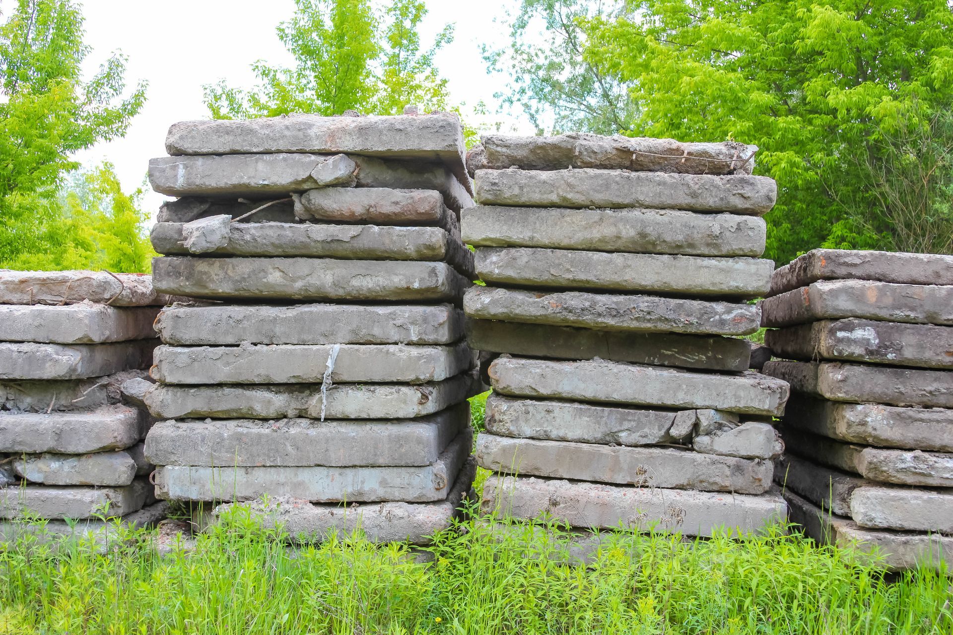 Stacks of gray concrete slabs piled outdoors in tall columns, with green grass and trees in the background