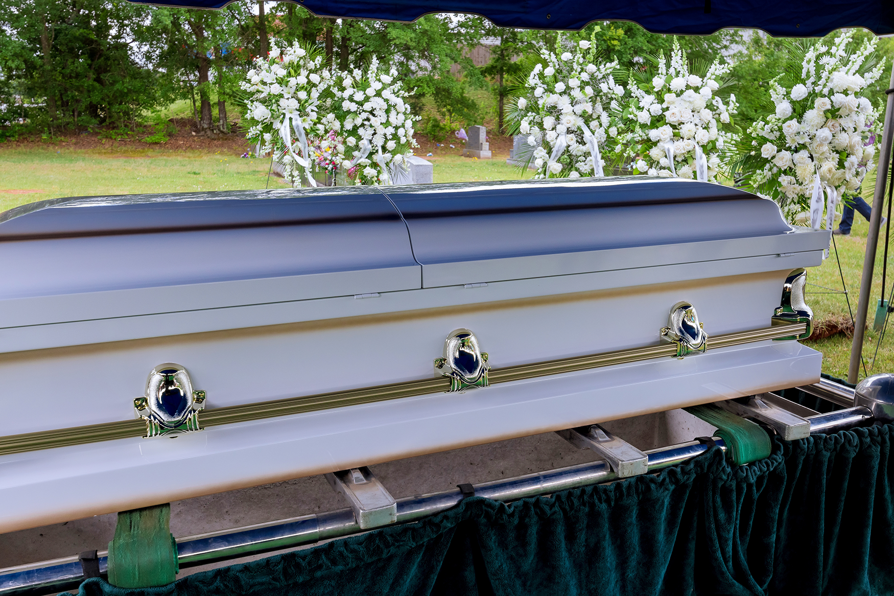 White coffin with green and gold accents at an outdoor funeral, surrounded by white flower arrangements.