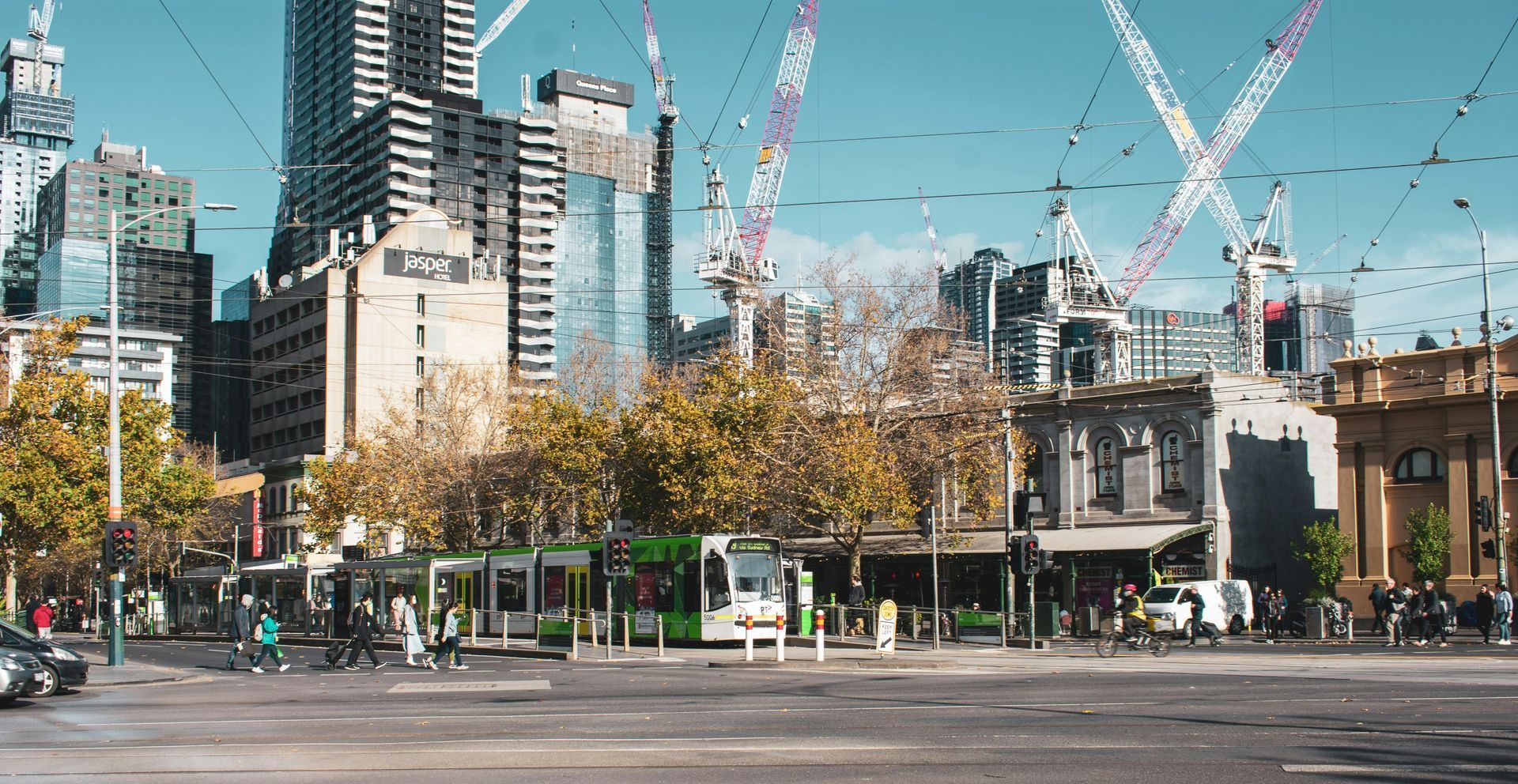 City street scene with tram, pedestrians, buildings, and construction cranes under a bright blue sky.