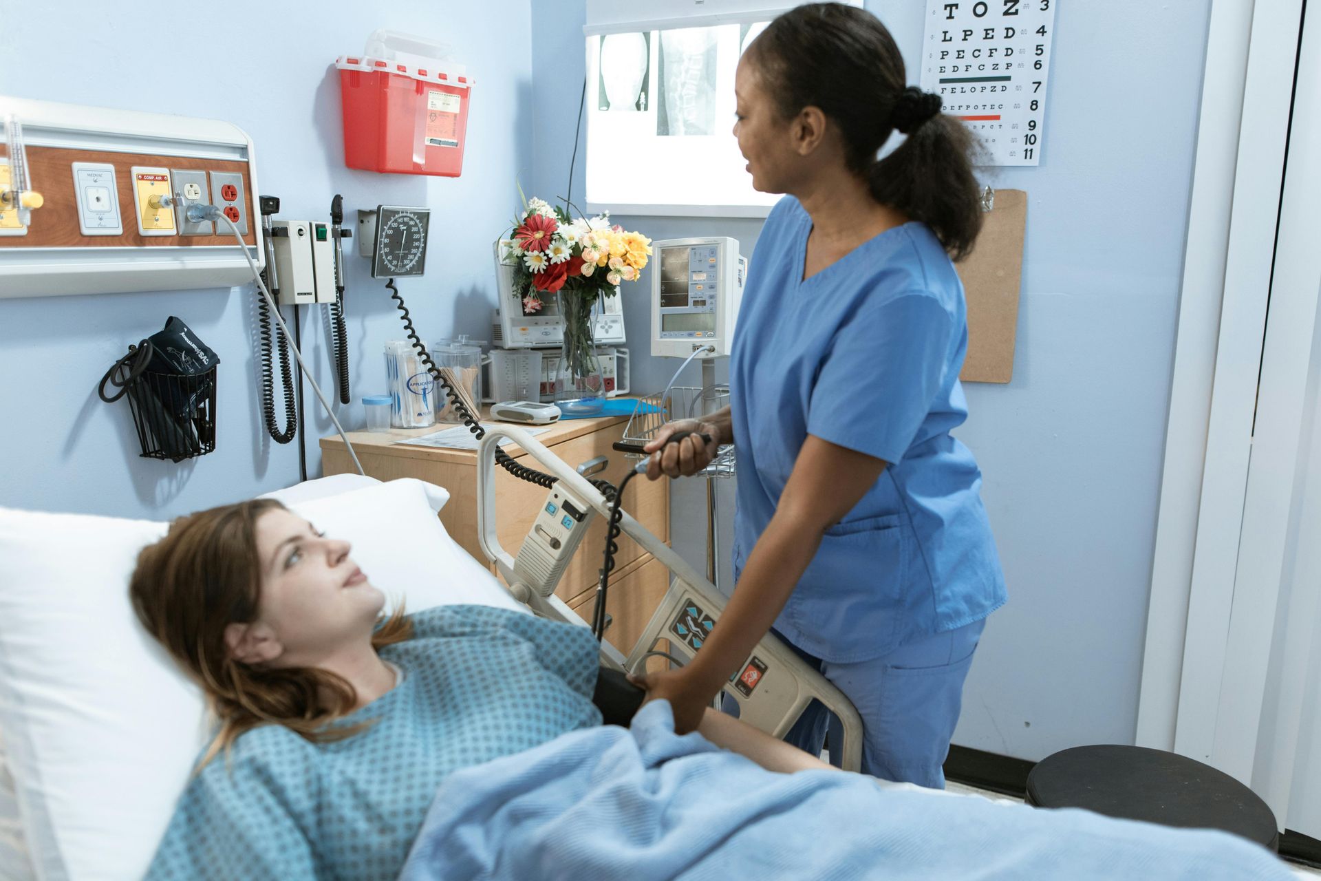 Nurse taking patient's blood pressure in a hospital room. Patient in bed, looking up. Flowers on a table.