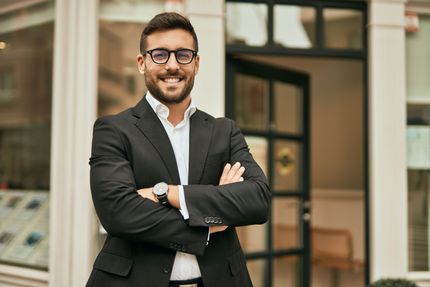 A man in a suit and glasses is standing in front of a building with his arms crossed.