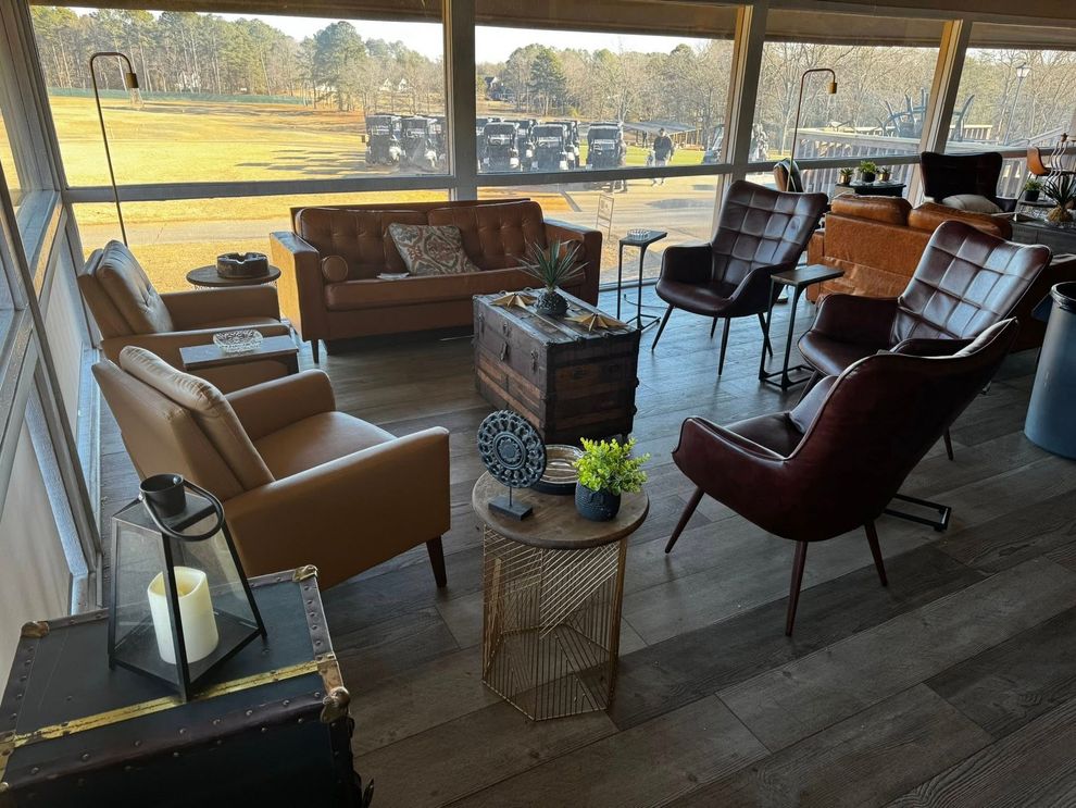 Cozy outdoor seating area with leather chairs, sofa, and small tables with decorative items. Large windows overlook a field.