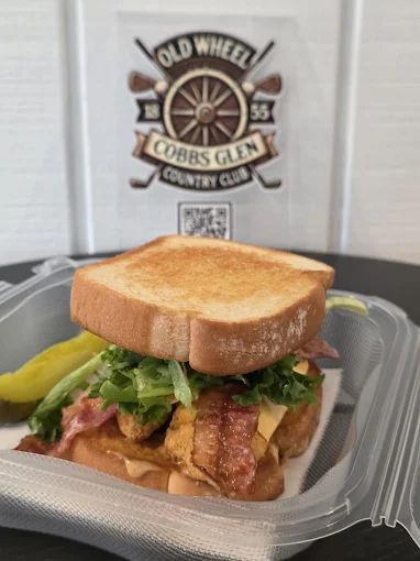 Sandwich with bacon, lettuce, and pickles in a takeout container in front of a Cobbs Glen Country Club logo.