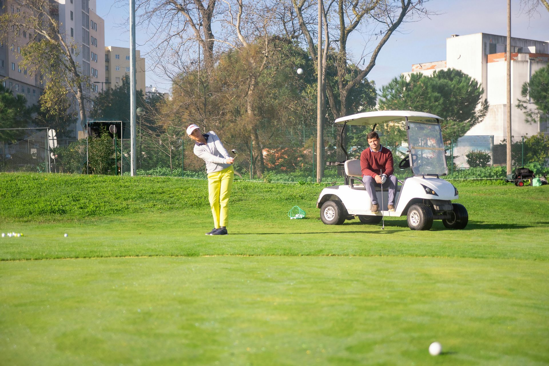 man hitting golf ball while another man watches from a golf cart nearby