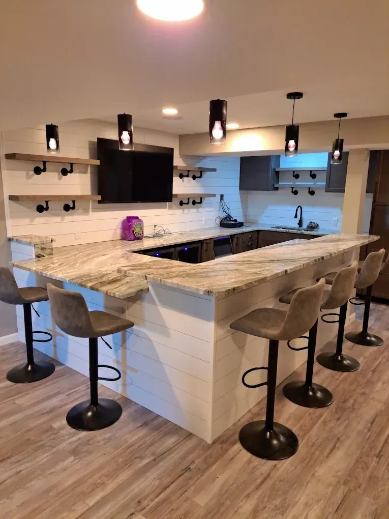 A modern basement bar features a U-shaped granite countertop, white shiplap base, and four grey bar stools on light wood floors.