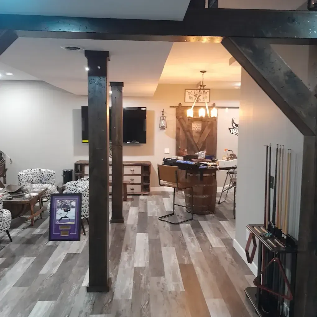 A spacious basement with rustic wood beams, gray flooring, a seating area with patterned chairs, and a bar with stools.