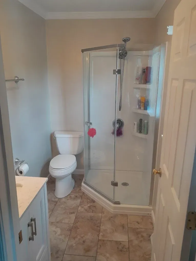 A neutral-toned bathroom with a corner glass shower, a white toilet, and a vanity cabinet on a tiled floor.
