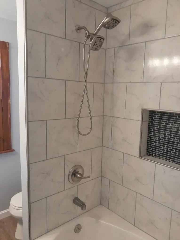 A shower and bathtub unit featuring white marbled tile walls, a dual shower head, and a recessed black tiled niche.