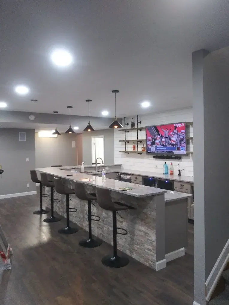 A basement bar with a stone-front counter, four bar stools, pendant lighting, floating shelves, and a wall-mounted TV.