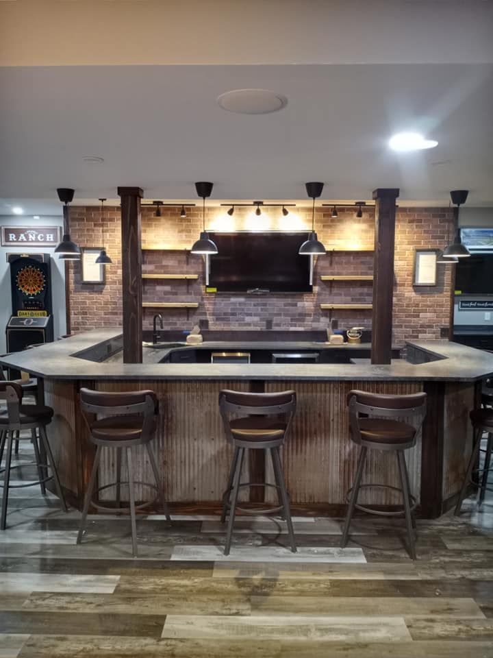 A rustic home bar with a concrete countertop, wood paneling, brick accent wall, mounted television, and three bar stools.