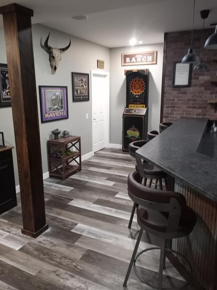 A finished basement home bar with gray wood floors, a dark counter, stools, and a dart board on a brick accent wall.