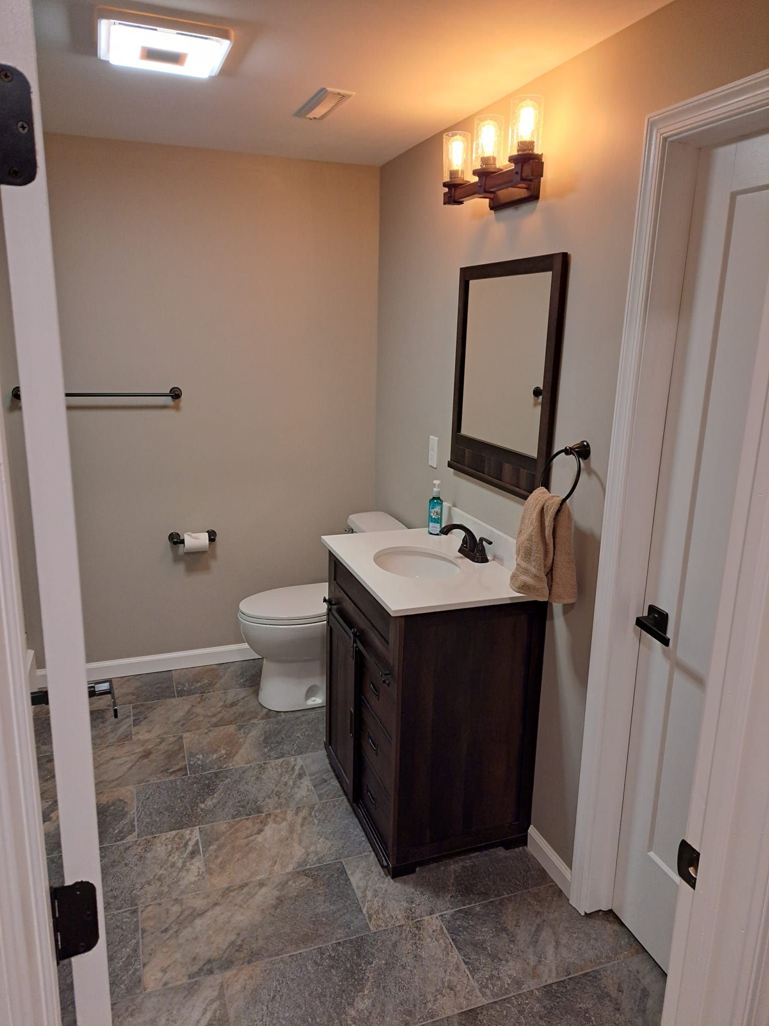 A half-bathroom with beige walls, a dark wood vanity, a rectangular mirror, overhead lighting, and stone-look flooring.