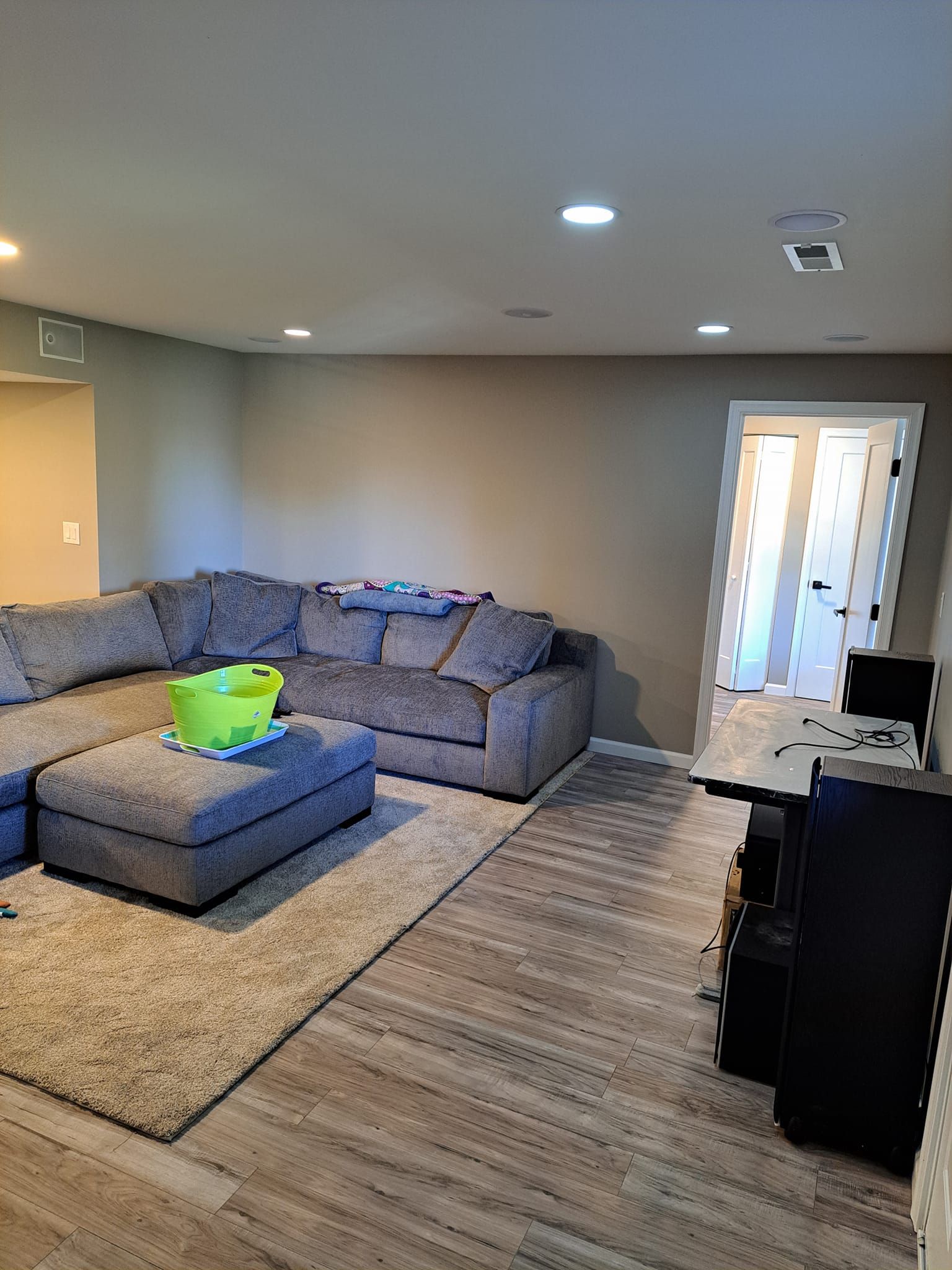 A gray sectional sofa sits on a rug in a neutrally painted room, near a doorway and a dark storage cabinet.