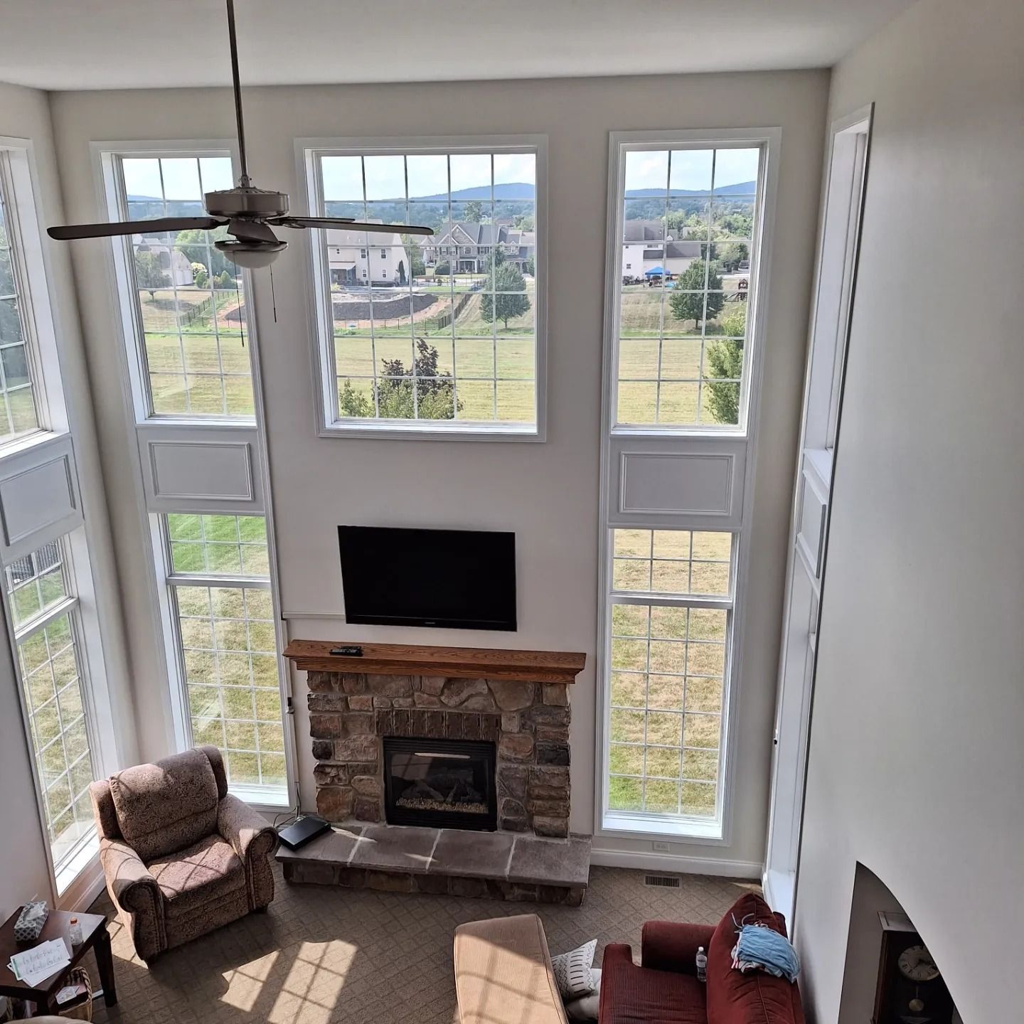 A high-angle view of a living room with a tall stone fireplace, a wall-mounted TV, and large windows looking onto a yard.