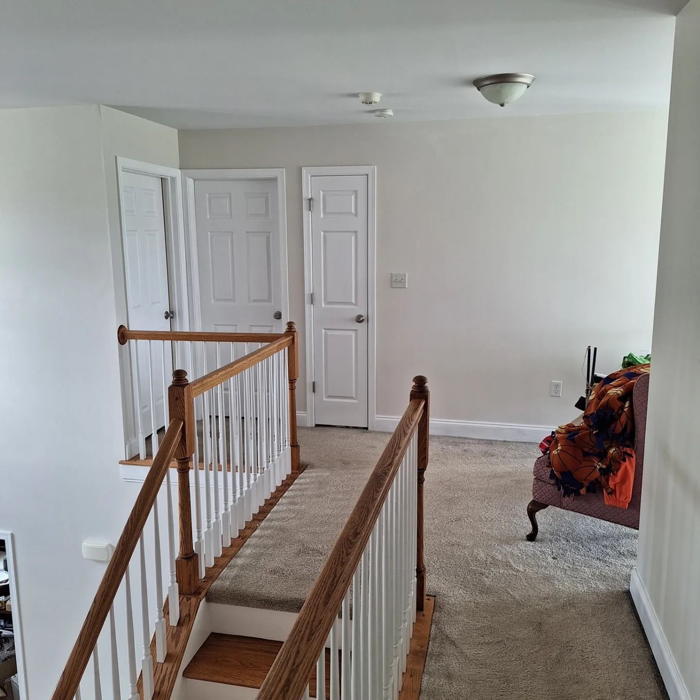 A carpeted landing with a wooden banister overlooking stairs, featuring three white doors and an armchair in the corner.