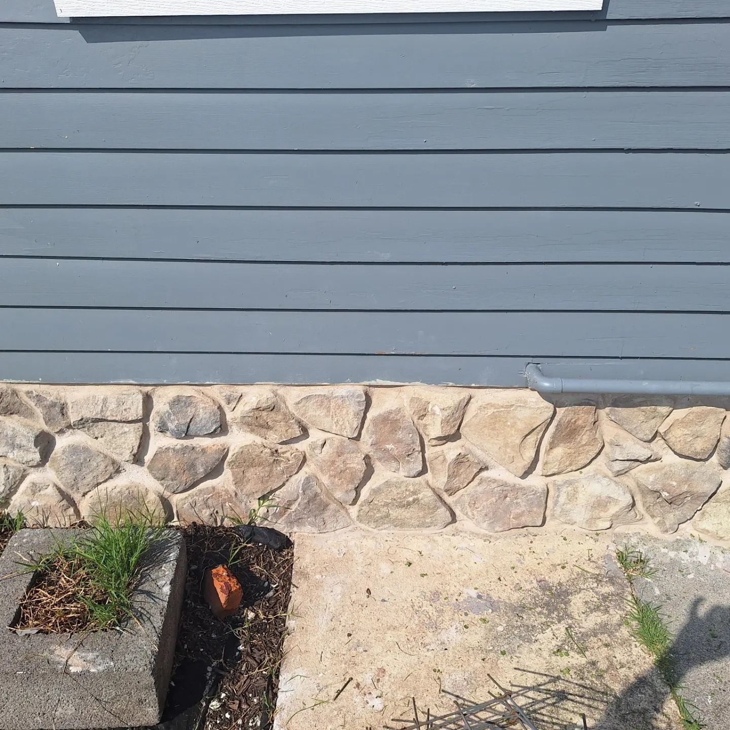 A light gray house siding sits above a foundation made of tan, rustic, irregular stones, next to a small garden planter.