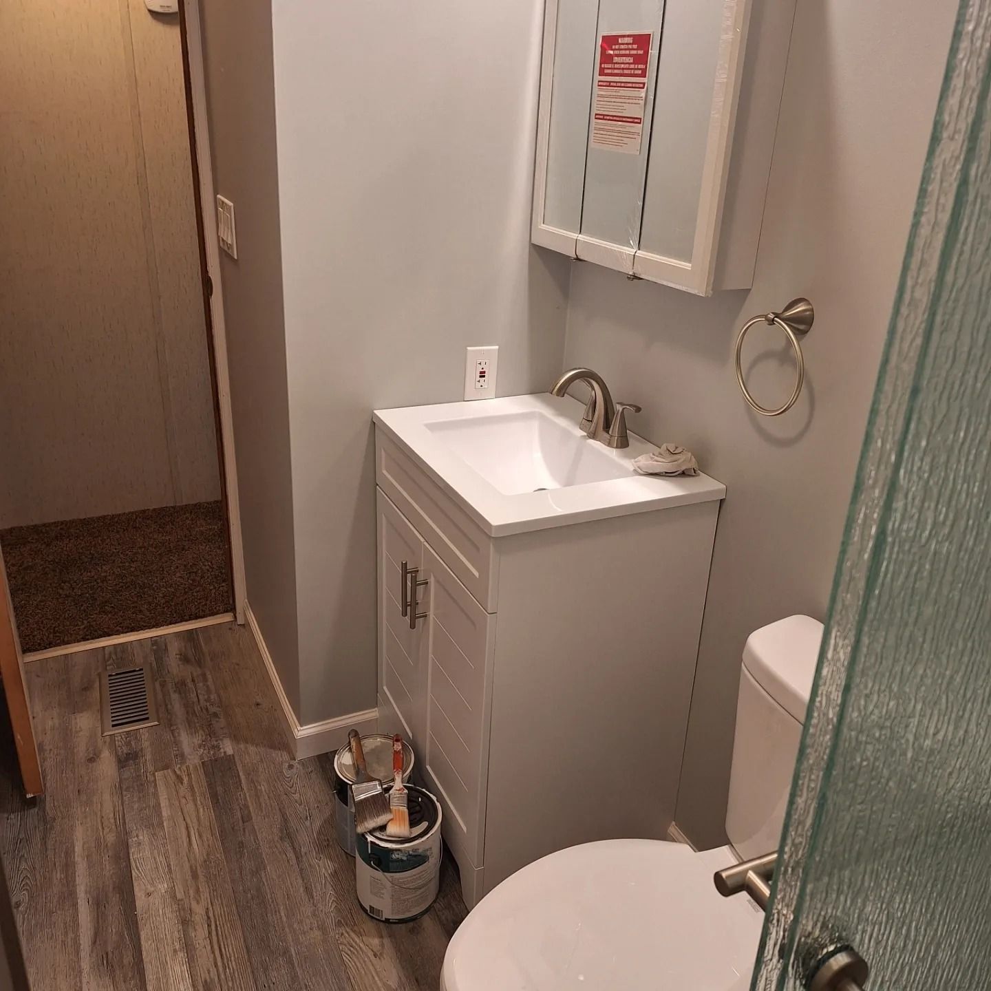 A small bathroom with light gray walls, white vanity, silver faucet, toilet, and a floor vent on wood-look flooring.