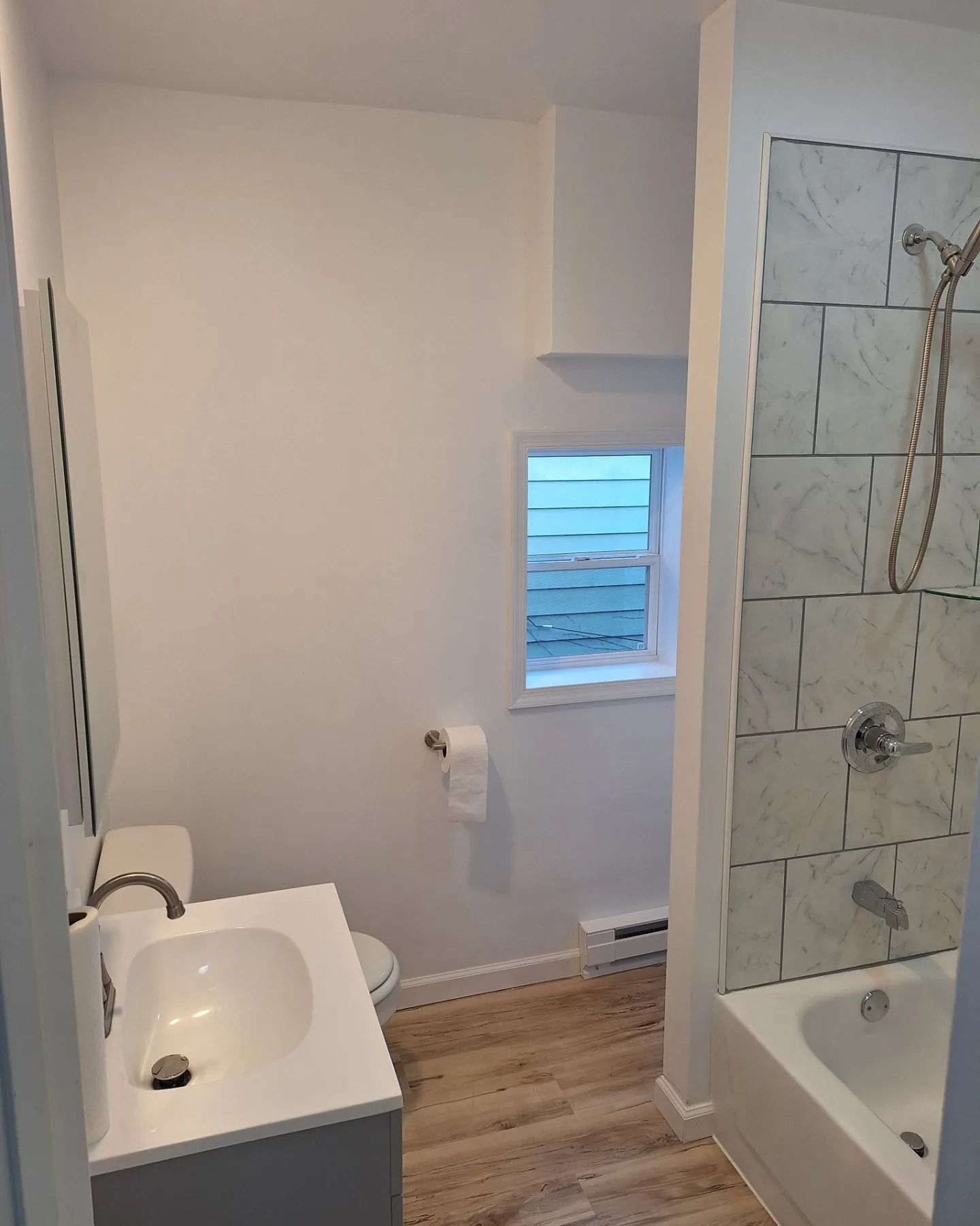 A bright bathroom featuring a white vanity with a silver faucet, a toilet, a window, and a bathtub with marble-look tiles.