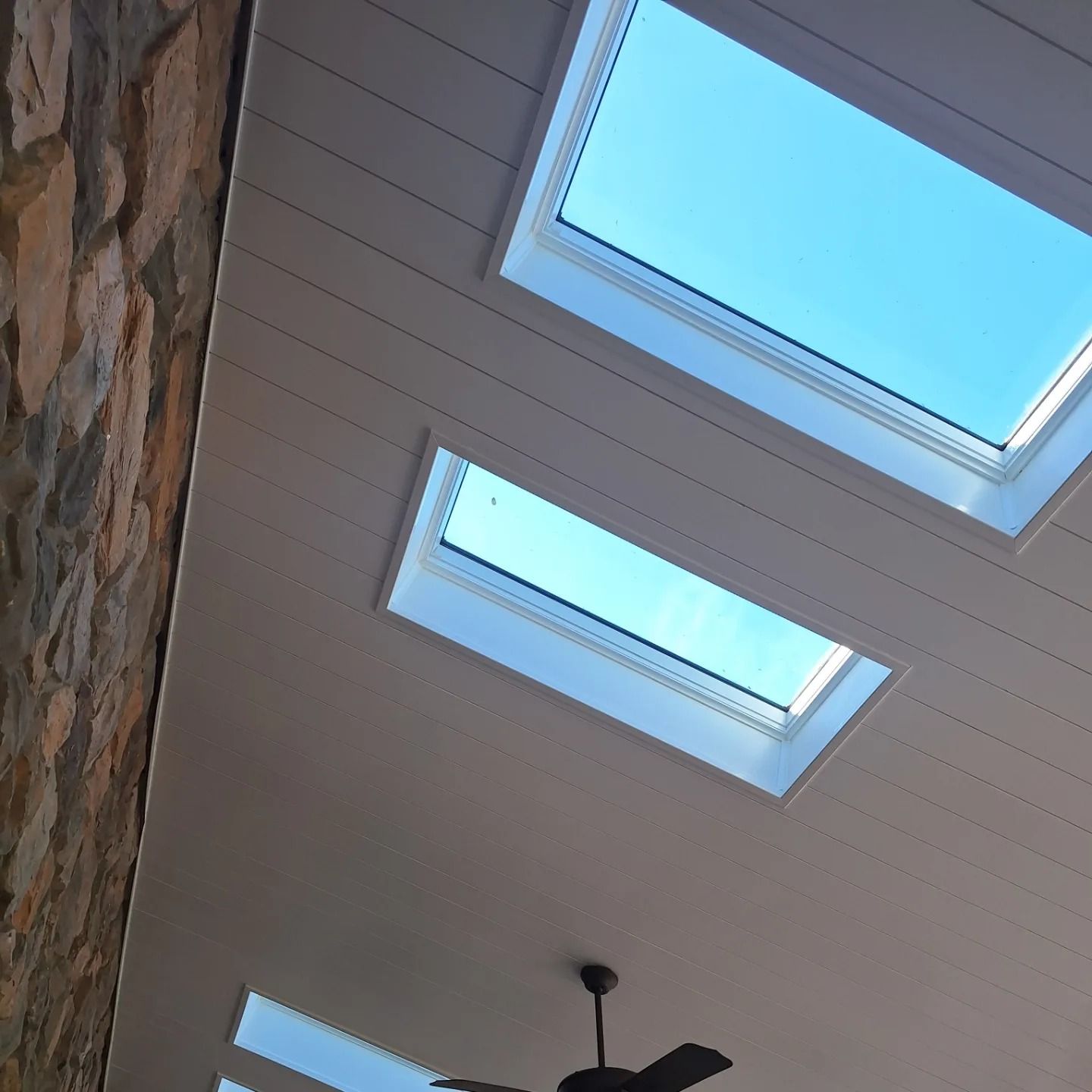 Low-angle view of a white paneled ceiling featuring three rectangular skylights and a ceiling fan against a stone wall.