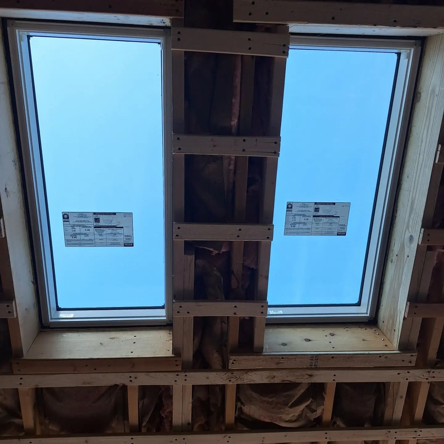 Two skylights framed by wooden beams against a clear blue sky, showing unfinished interior construction.