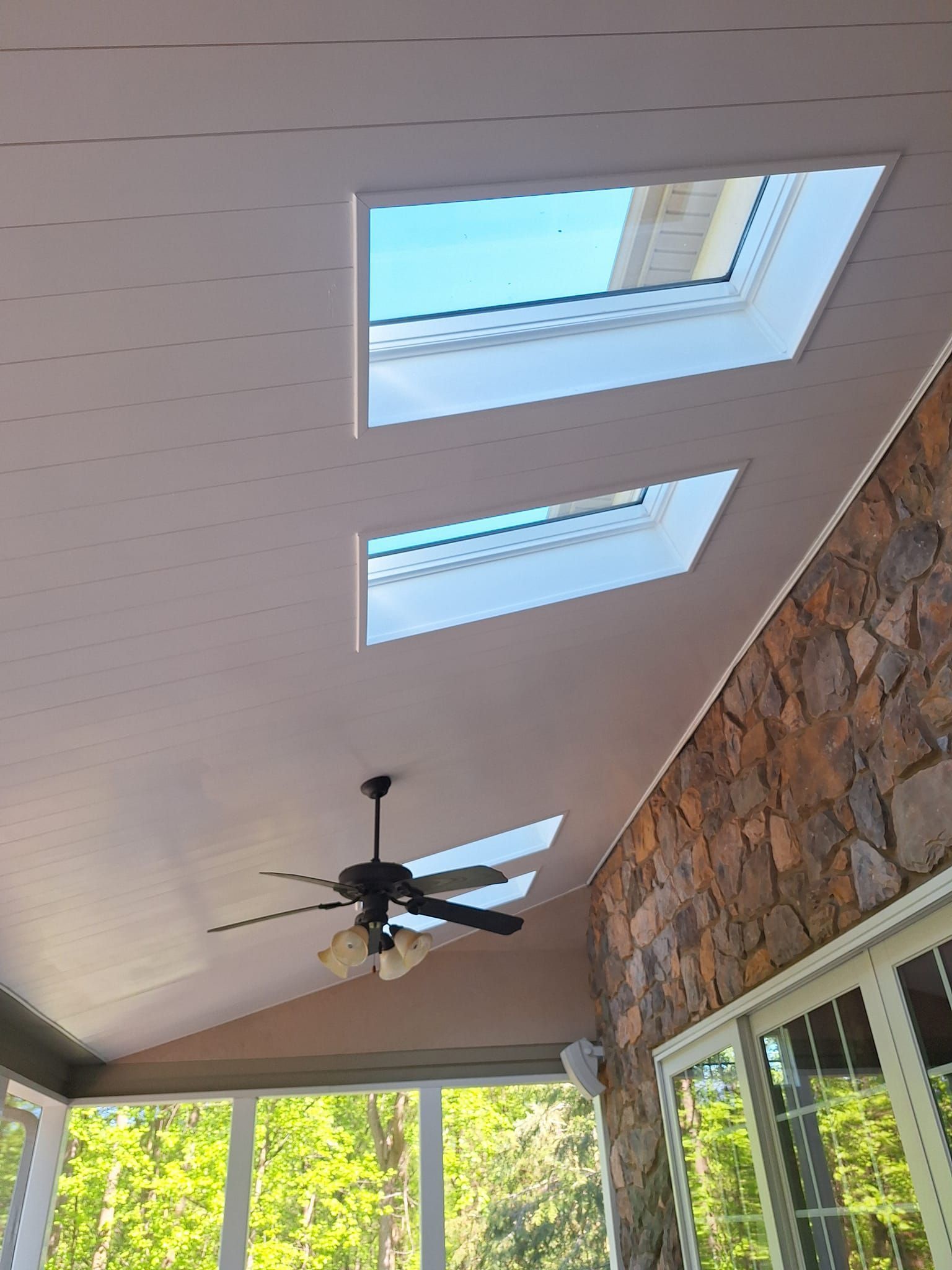 Sunroom ceiling with white tongue-and-groove boards, two rectangular skylights, a ceiling fan, and stone accent wall.