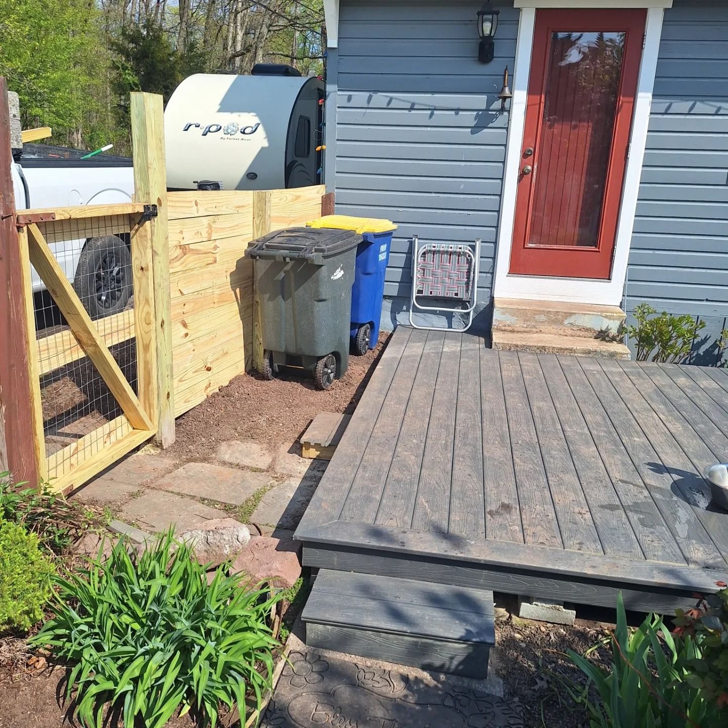 A wooden deck and gate area outside a house, with two trash cans next to a small trailer parked in the yard.