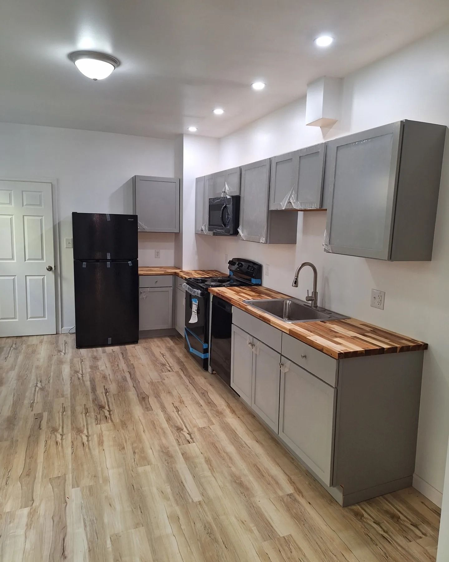 A galley-style kitchen with grey cabinets, wood countertops, a black refrigerator, and light wood-look flooring.