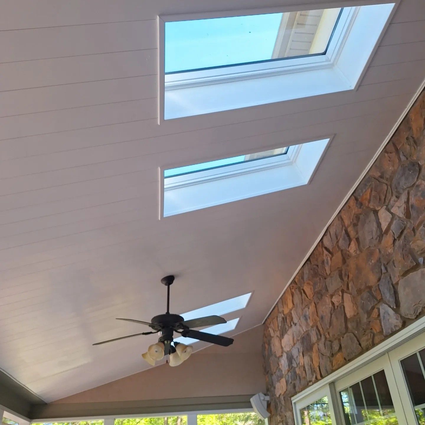 A white sloped ceiling with two skylights, a ceiling fan, and a rustic stone wall in a bright, indoor living space.