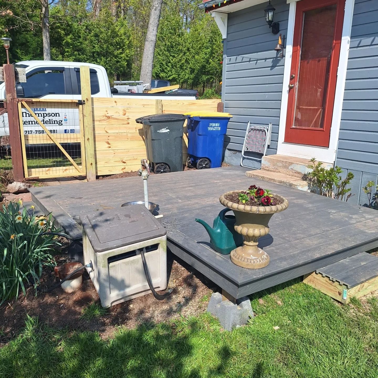A gray patio deck with a dark blue and a black trash bin, a hose reel, and a decorative planter outside a house.