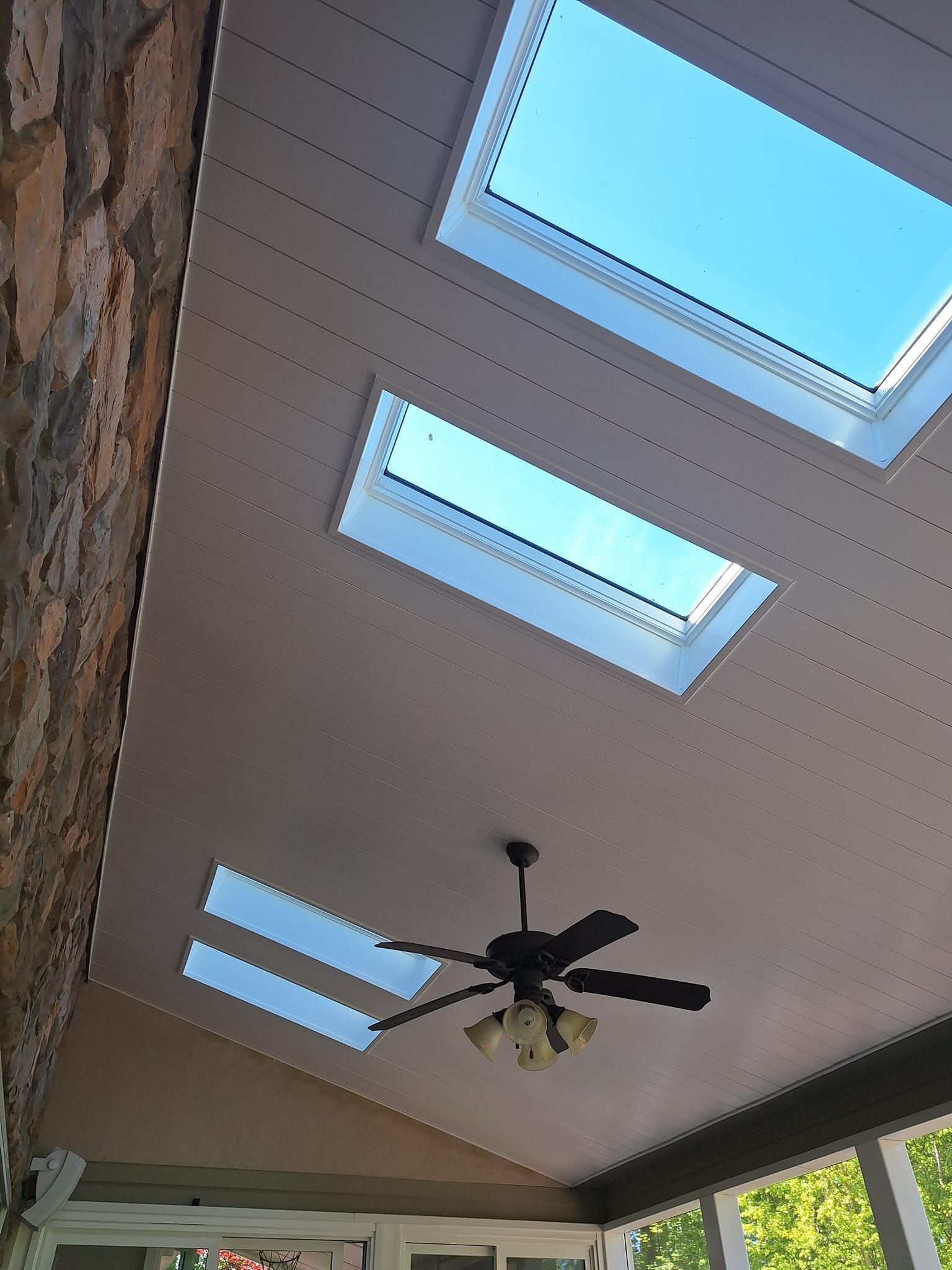 A sloped wooden ceiling with three rectangular skylights and a hanging ceiling fan in a sunroom.