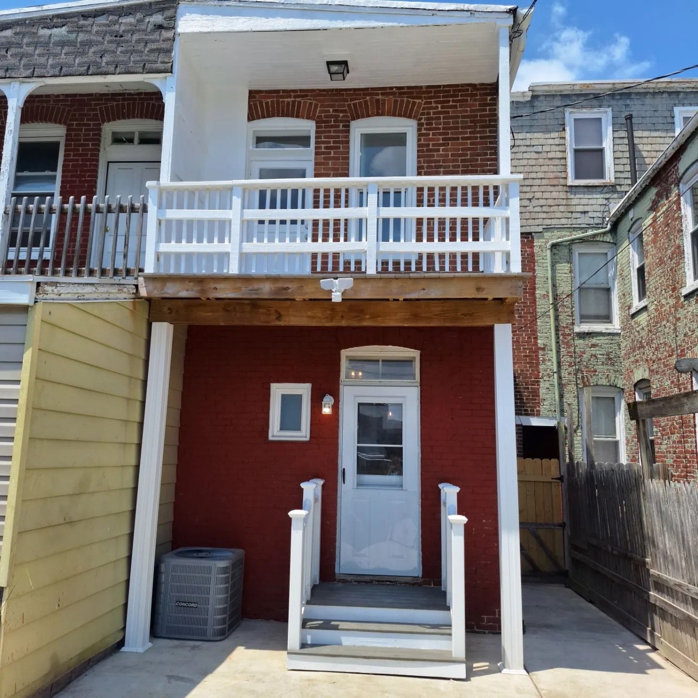 A red brick townhouse with a white wooden balcony, stairs leading to the entrance, and a side view of neighboring homes.