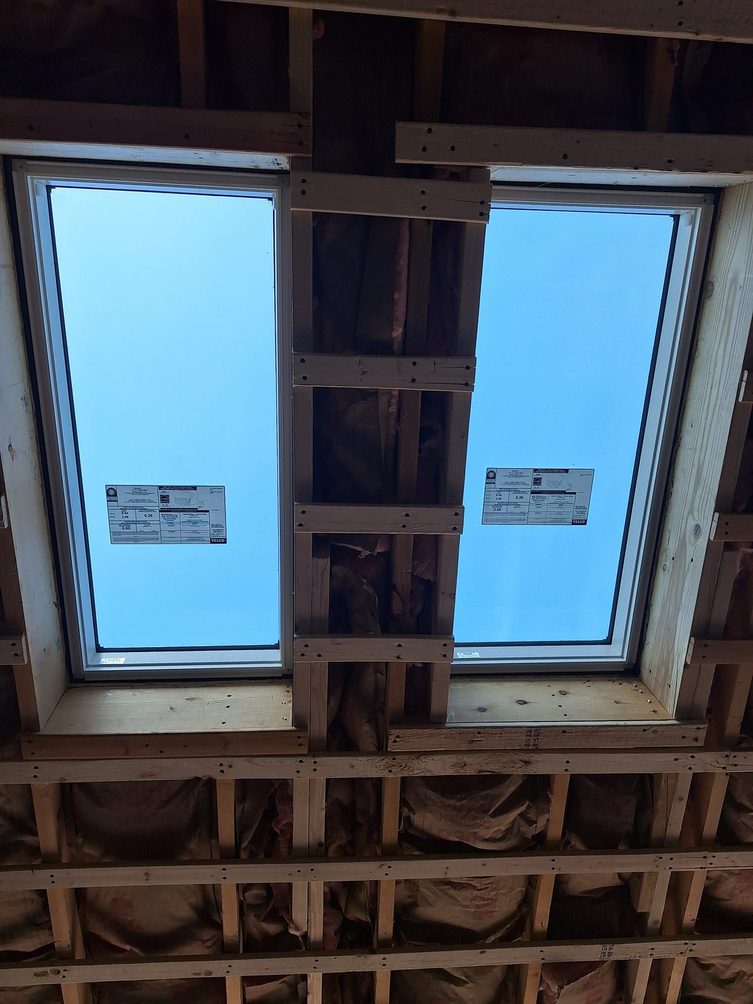 Two skylights installed in a wooden attic frame with exposed insulation and rafters against a clear blue sky.