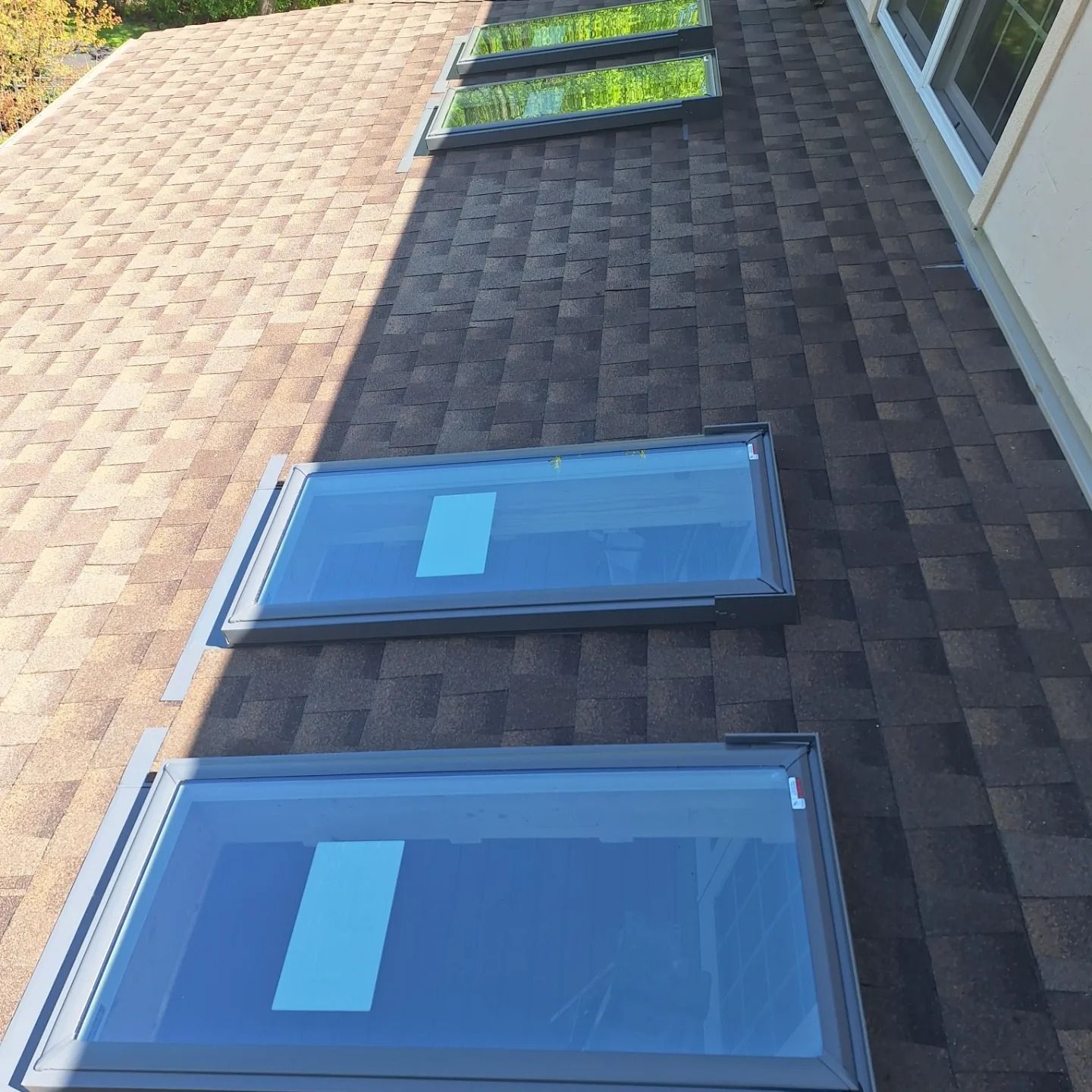 Four skylights installed in a dark brown shingled roof next to a house wall.