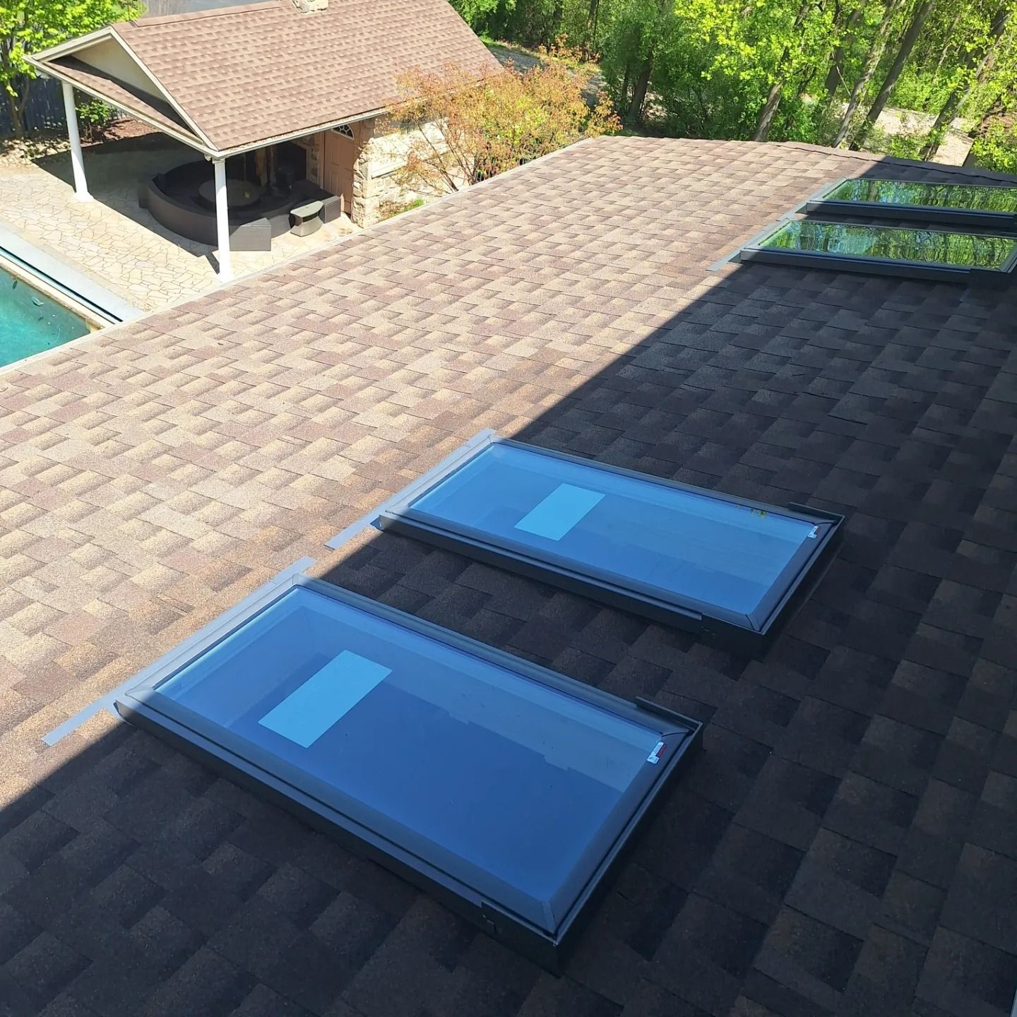 A view of a dark shingled roof with several installed skylights, overlooking a backyard with a pool and a gazebo.