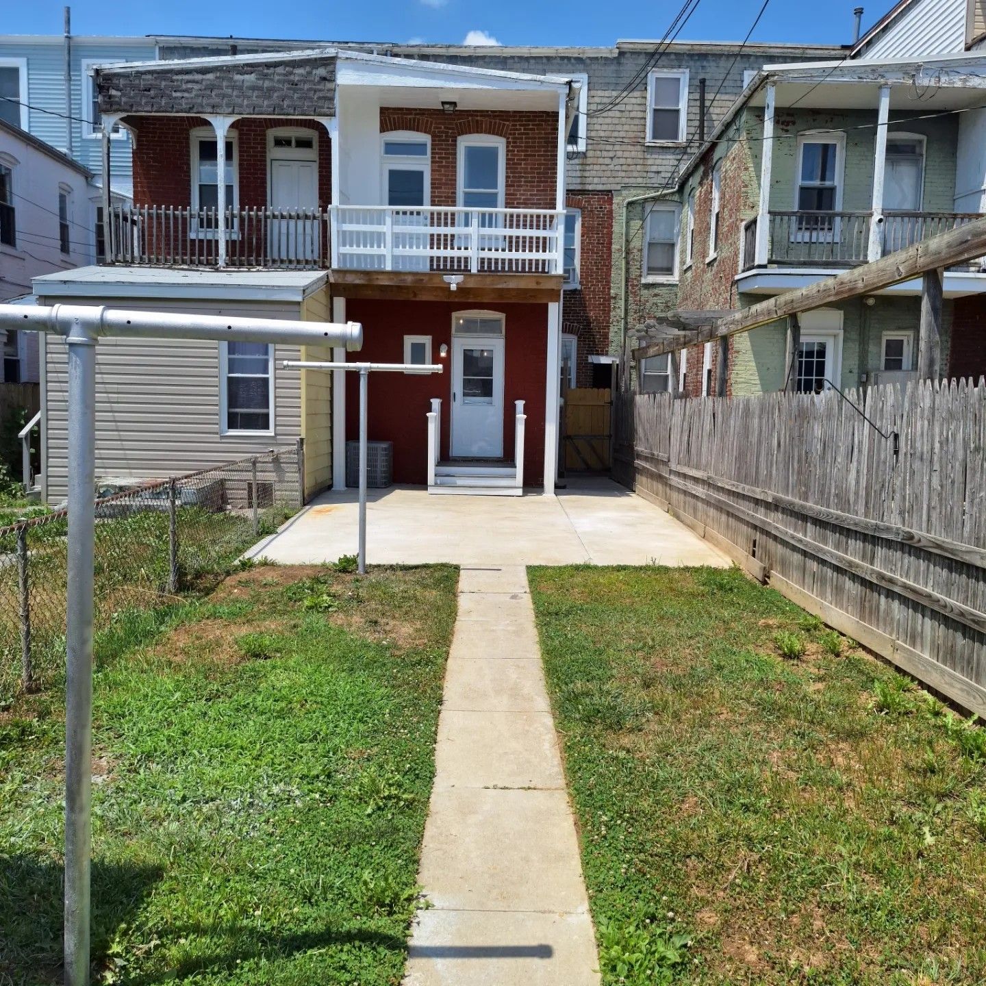 A backyard with a concrete patio, a central walkway, and a brick row house with a white wooden balcony and stairs.