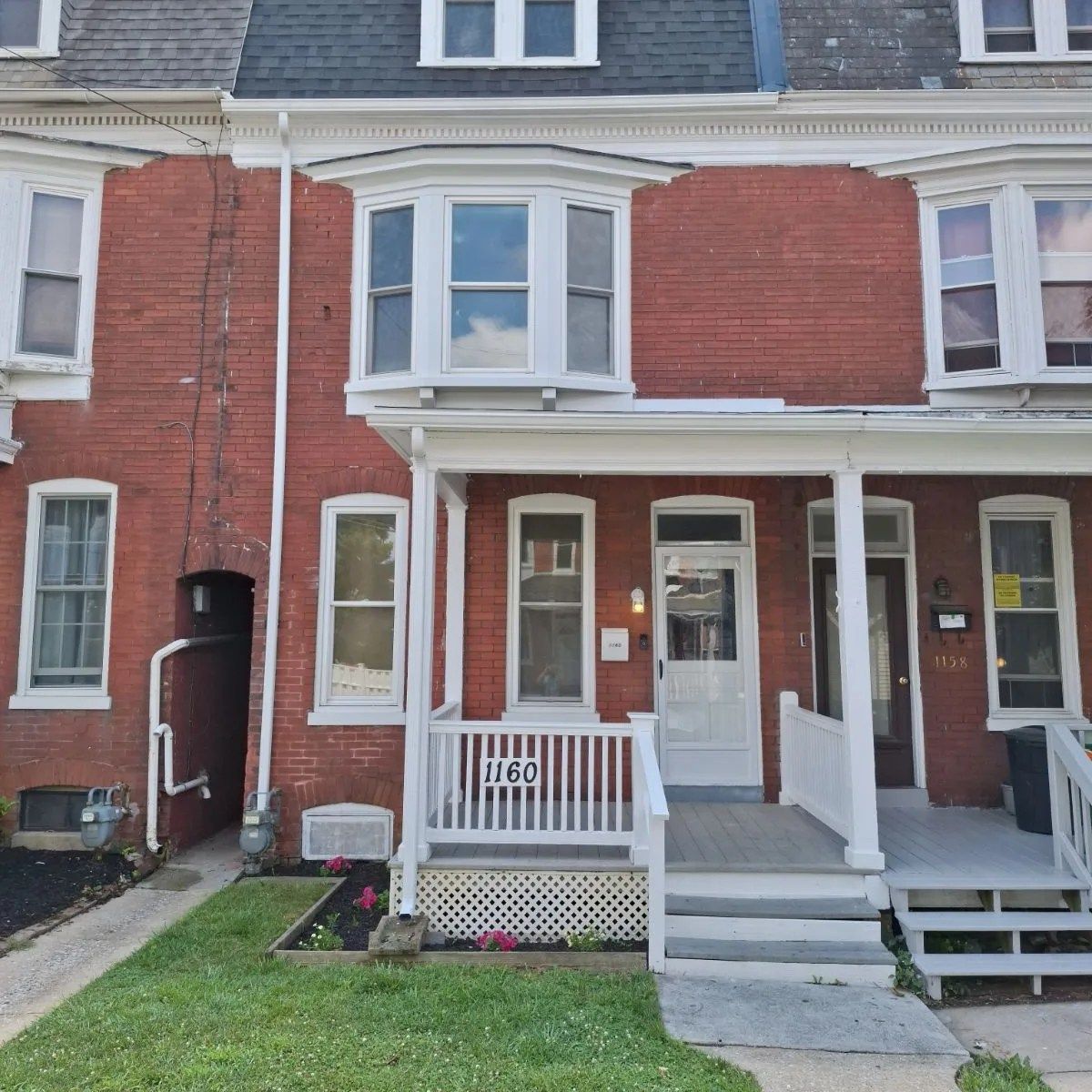 A red brick row house with a white front porch, bay window on the second floor, and white trim on a sunny day.