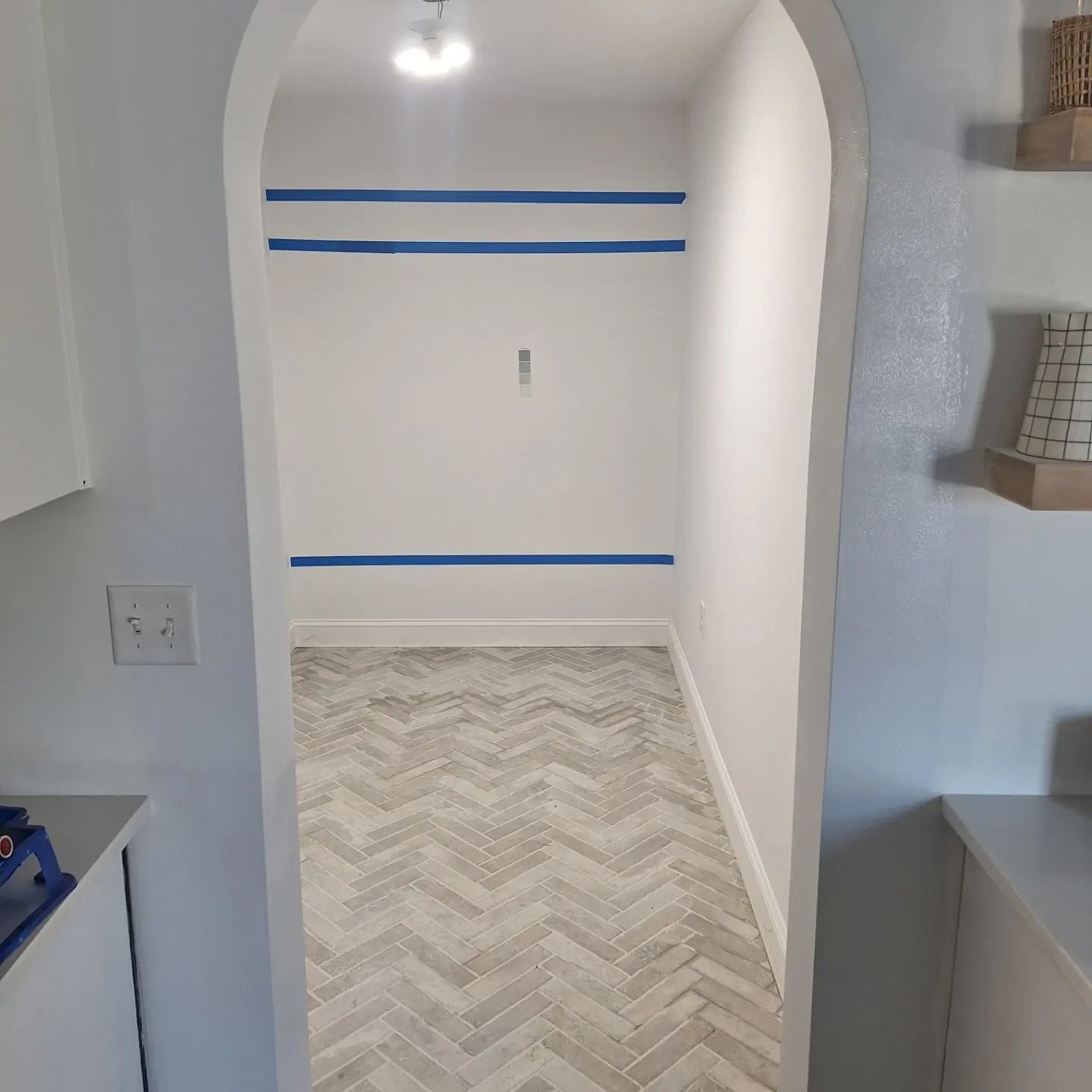 An arched doorway leads to a small room with herringbone patterned floor tiles and three strips of blue painter's tape.
