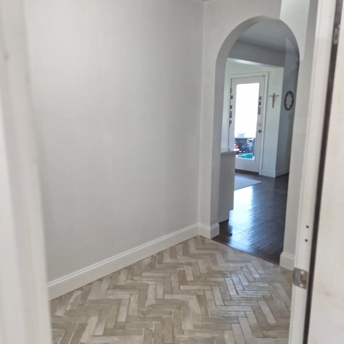 A light gray room with light-colored herringbone patterned flooring leading through an arched doorway to another room.