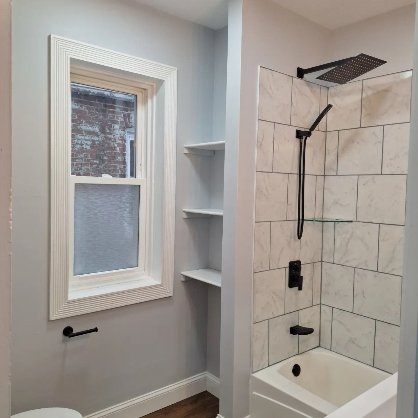A bathroom featuring a white-trimmed window, built-in shelving, and a tiled shower with matte black fixtures.