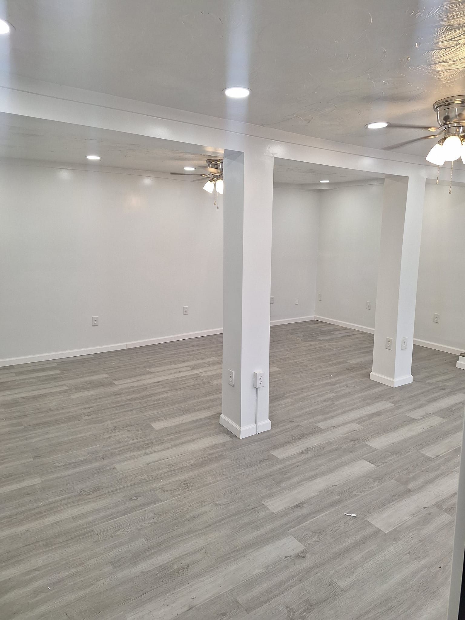 A bright, empty room with light gray wood-look flooring, white walls, two white support columns, and ceiling lights.