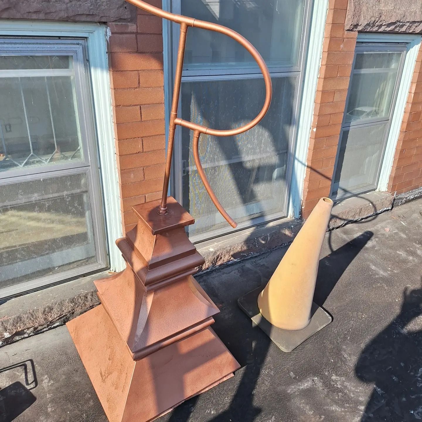 A copper finial with a decorative curved metal rod sits on a dark rooftop next to a small, tan traffic cone.