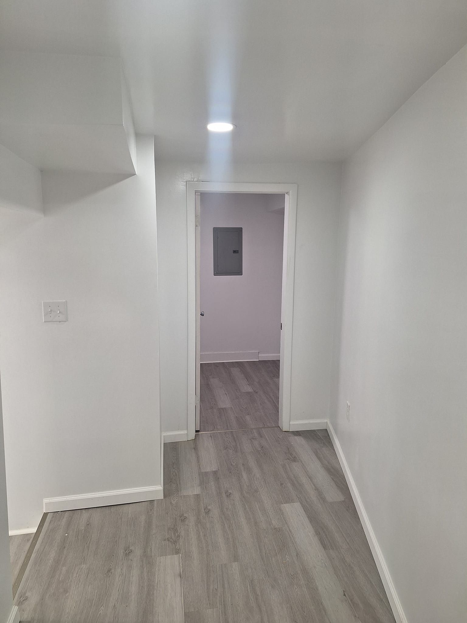 A view down a bright, narrow hallway with white walls, grey wood-look flooring, and a doorway leading to another room.